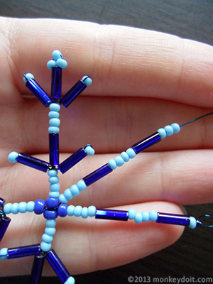 Wrap the wire around the centre of the snowflake and come out in between two of its arms. String the following sequence of beads onto the wire: four small seed beads, one bugle bead, four small seed beads, one bugle bead and three small seed beads